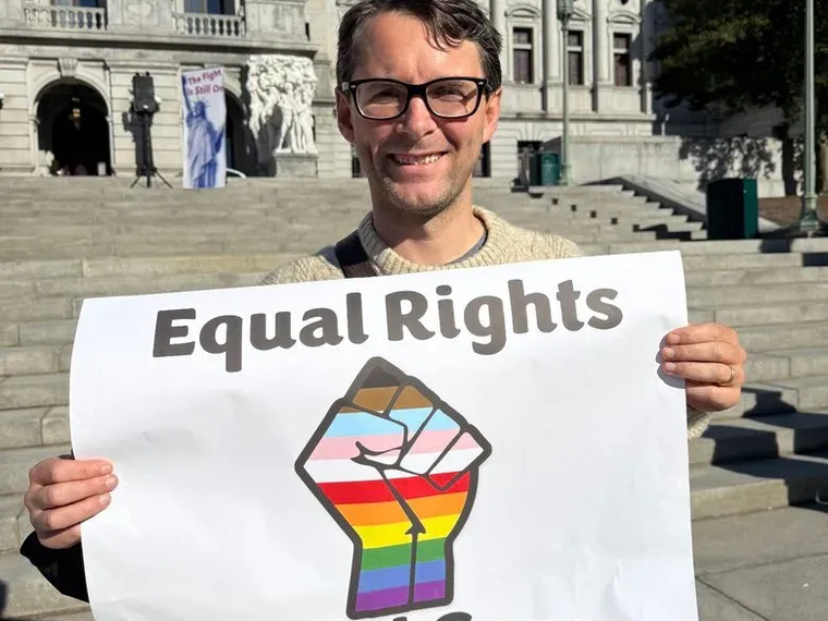 Tom holding a pro trans rights placard saying "Equal Rights Equal Care"
